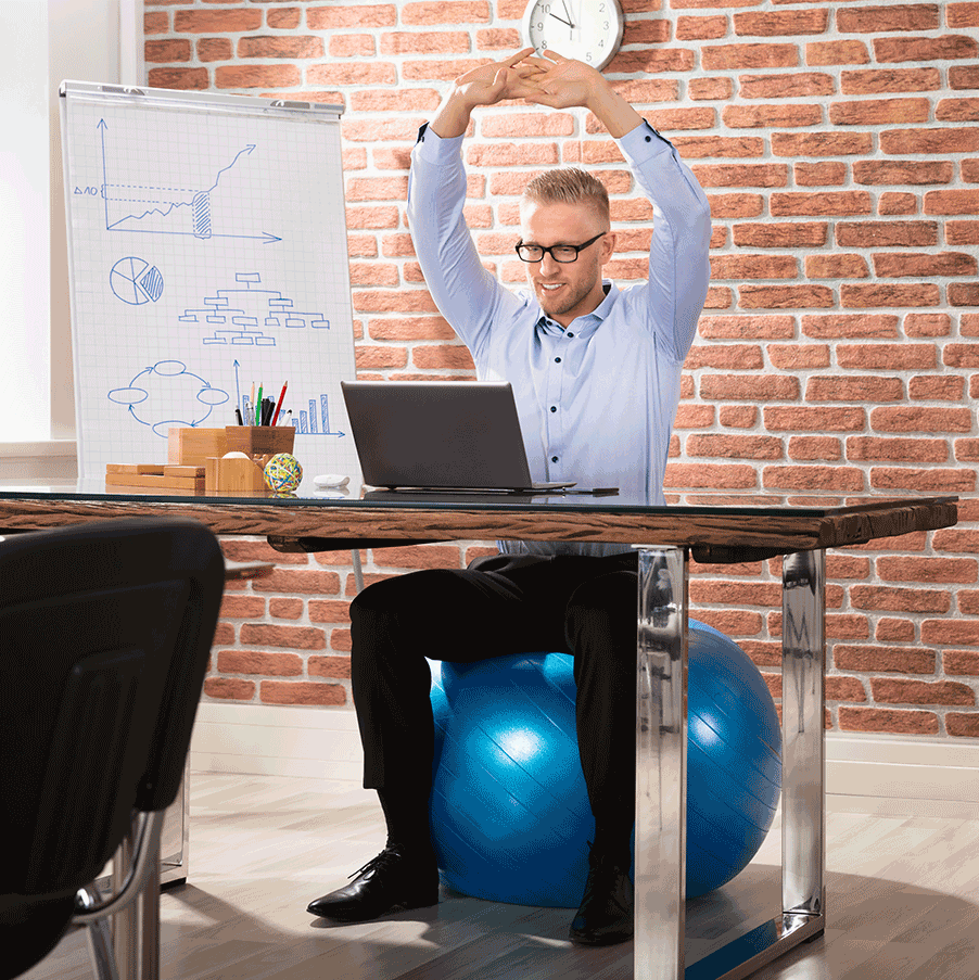 Person in Businesskleidung streckt die Arme über den Kopf, während sie auf einem Fitnessball an einem Schreibtisch mit Laptop sitzt, was auf einen Online-Firmenfitnesskurs hinweist. Im Hintergrund zeigt ein Whiteboard verschiedene Diagramme und Grafiken, die den Fokus auf Produktivität und Gesundheitsmetriken legen.